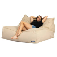 Woman relaxing on a beige bean bag chair with a visible brand logo.