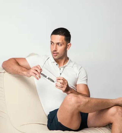 Man sitting on a couch wearing a white polo shirt and dark shorts, holding glasses.