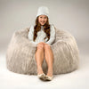 Young girl sitting on a fluffy gray bean bag chair wearing a white sweater and hat.