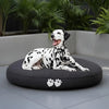 Dalmatian dog lying on a black round dog bed with paw prints in an outdoor setting.