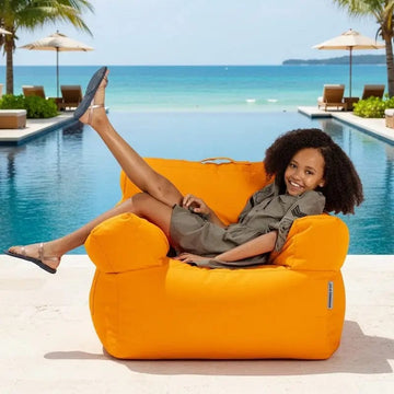 Woman relaxing on an orange bean bag chair by a pool with a beach in the background